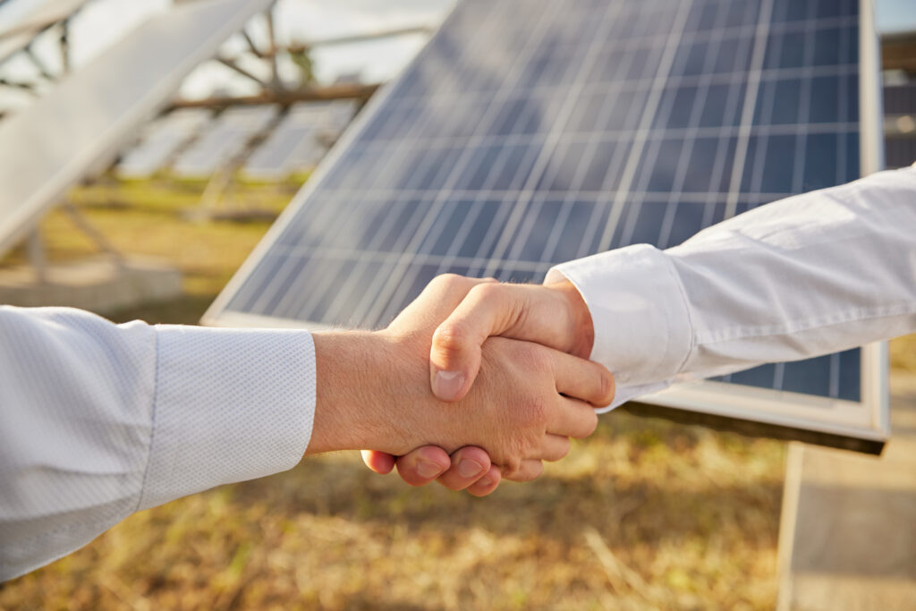 Crop anonymous male business partners shaking hands as symbol of agreement while standing near photovoltaic panels on agricultural solar power station